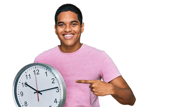 Young handsome hispanic man holding big clock smiling happy pointing with hand and finger