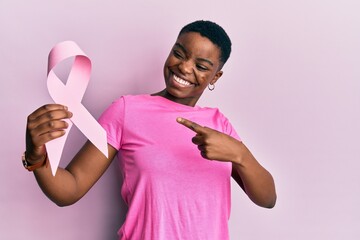 Young african american woman holding pink cancer ribbon smiling happy pointing with hand and finger