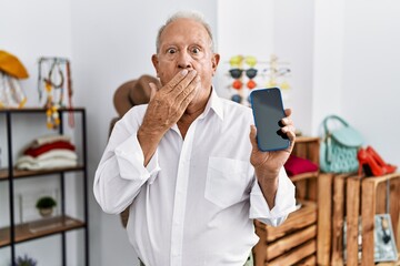 Senior man holding smartphone at retail shop covering mouth with hand, shocked and afraid for mistake. surprised expression