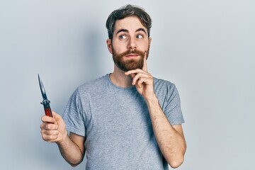 Caucasian man with beard holding pocket knife serious face thinking about question with hand on...