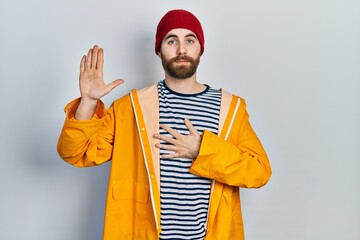 Caucasian man with beard wearing yellow raincoat swearing with hand on chest and open palm, making...