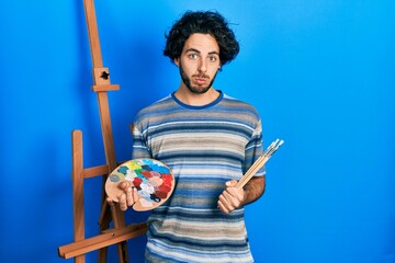 Handsome hispanic man standing drawing with palette by painter easel stand depressed and worry for...