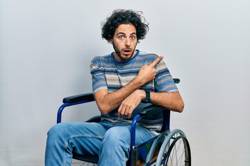 Handsome hispanic man sitting on wheelchair surprised pointing with finger to the side, open mouth...