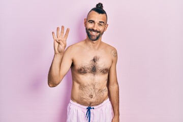 Fototapeta premium Young hispanic man shirtless wearing swimsuit showing and pointing up with fingers number four while smiling confident and happy.