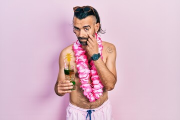 Young hispanic man wearing swimsuit and hawaiian lei drinking tropical cocktail pointing to the eye...