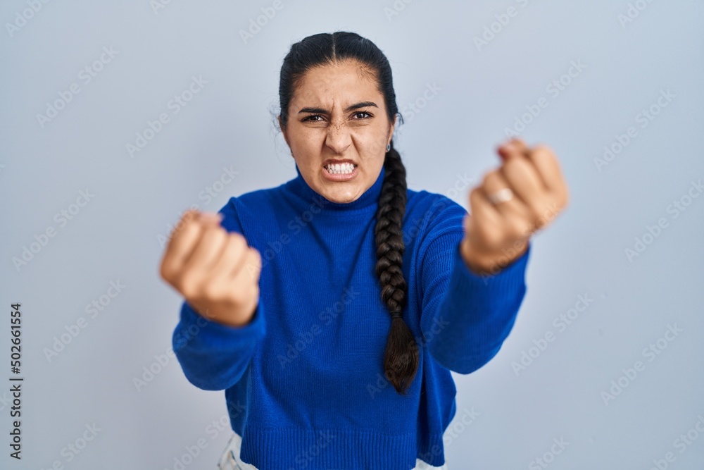 Poster Young hispanic woman standing over isolated background angry and mad raising fists frustrated and furious while shouting with anger. rage and aggressive concept.