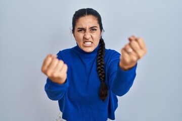 Young hispanic woman standing over isolated background angry and mad raising fists frustrated and furious while shouting with anger. rage and aggressive concept.