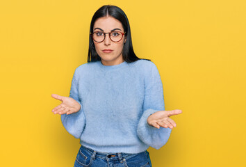Young hispanic woman wearing casual clothes and glasses clueless and confused expression with arms and hands raised. doubt concept. - obrazy, fototapety, plakaty