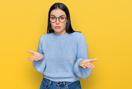 Young hispanic woman wearing casual clothes and glasses clueless and confused expression with arms and hands raised. doubt concept.