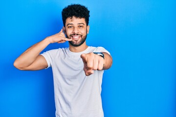 Young arab man with beard wearing casual white t shirt smiling doing talking on the telephone gesture and pointing to you. call me.