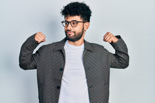 Young arab man with beard wearing glasses showing arms muscles smiling proud. fitness concept.