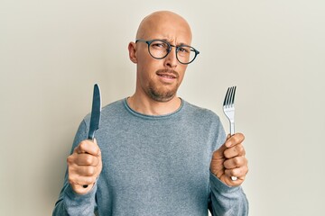 Bald man with beard holding fork and knife ready to eat clueless and confused expression. doubt...