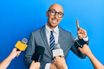 Bald man with beard being interviewed by reporters holding microphones smiling with an idea or question pointing finger up with happy face, number one