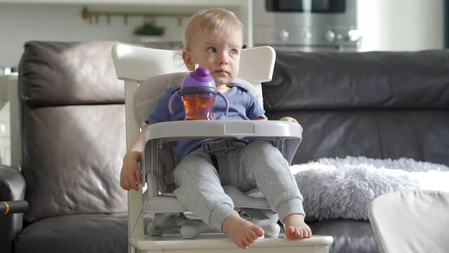 Cute kid with baby straw feeding cup sitting in booster seat, one year old toddler watching tv. High quality 4k footage