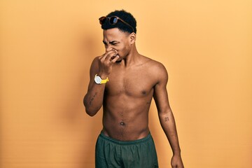Young african american man wearing swimwear and swimmer glasses smelling something stinky and...