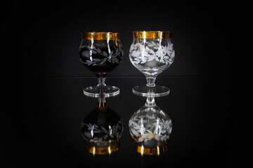 Two glasses - one with dark liquid and another empty on mirror black surface