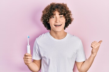 Handsome young man holding electric toothbrush pointing thumb up to the side smiling happy with open mouth
