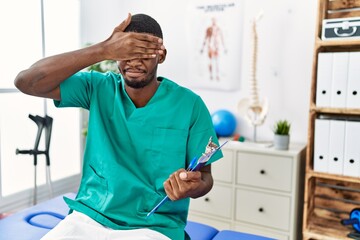Obraz premium Young african american man working at pain recovery clinic covering eyes with hand, looking serious and sad. sightless, hiding and rejection concept