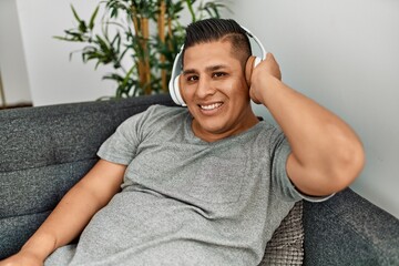 Young hispanic man smiling happy listening music sitting on the sofa at home.