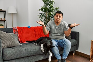 Young latin man and dog sitting on the sofa at home clueless and confused expression with arms and hands raised. doubt concept.