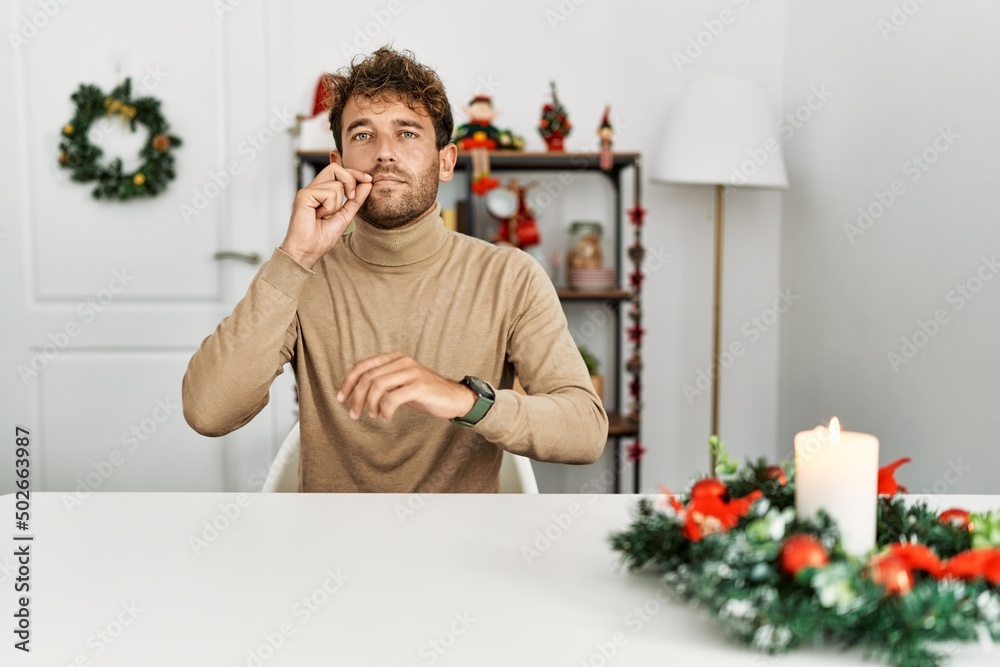 Wall mural young handsome man with beard sitting on the table by christmas decoration mouth and lips shut as zi