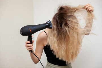 a girl with blond hair dries them with a hair dryer on a light background