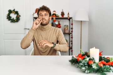 Young handsome man with beard sitting on the table by christmas decoration mouth and lips shut as...