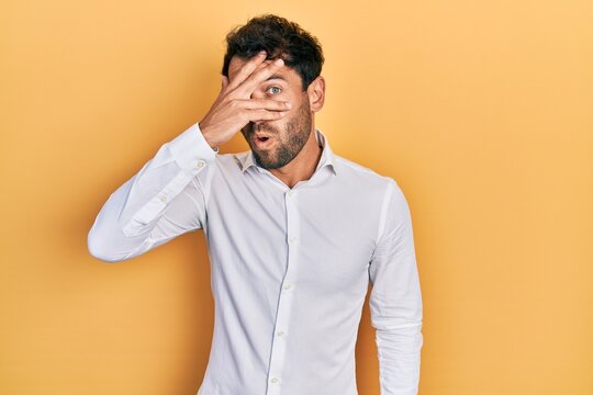 Handsome man with beard wearing casual white t shirt peeking in shock covering face and eyes with hand, looking through fingers afraid