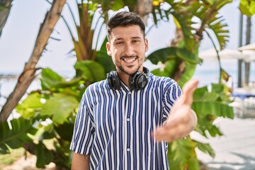 Young handsome man listening to music using headphones outdoors smiling friendly offering handshake as greeting and welcoming. successful business.