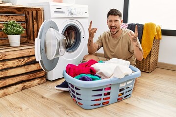 Young handsome man putting dirty laundry into washing machine shouting with crazy expression doing...