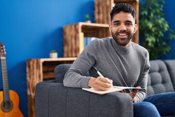 Young hispanic man writing on book sitting on sofa at home