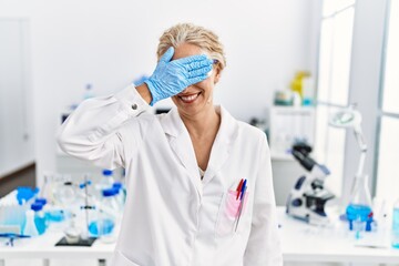 Middle age blonde woman working at scientist laboratory smiling and laughing with hand on face covering eyes for surprise. blind concept.