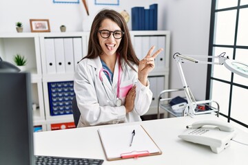 Young doctor woman wearing doctor uniform and stethoscope at the clinic smiling with happy face winking at the camera doing victory sign. number two.