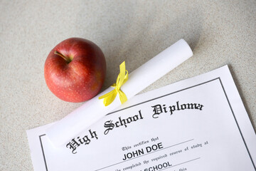 A high school diploma lies on table with small scroll and red apple. Education documents