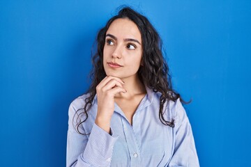Young brunette woman standing over blue background with hand on chin thinking about question,...