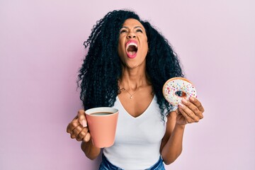 Middle age african american woman eating doughnut and drinking coffee angry and mad screaming...