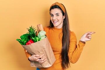 Young brunette teenager holding paper bag with groceries thinking concentrated about doubt with finger on chin and looking up wondering