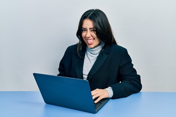 Beautiful hispanic woman working at the office with laptop sticking tongue out happy with funny...