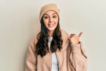 Young hispanic woman wearing wool sweater and winter hat smiling with happy face looking and pointing to the side with thumb up.