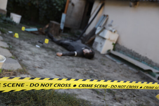 Victim of a violent crime in a backyard of residental house in evening. Dead man body under the yellow police line tape and evidence markers on crime scene