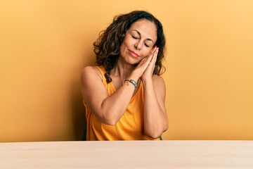 Beautiful middle age woman wearing casual clothes sitting on the table sleeping tired dreaming and posing with hands together while smiling with closed eyes.