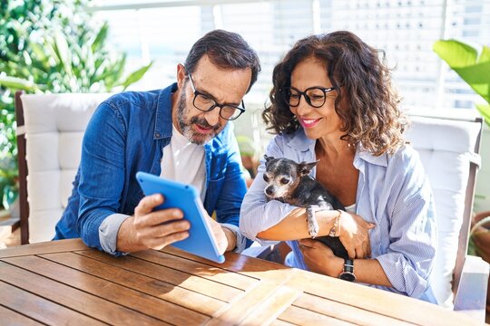 Middle age hispanic couple using touchpad sitting on table with dog at terrace