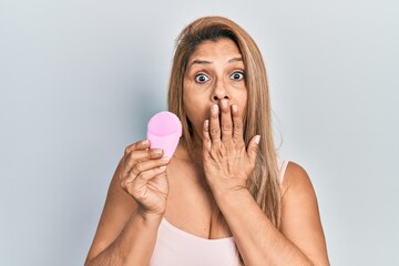 Middle age hispanic woman using facial exfoliating cleaner covering mouth with hand, shocked and afraid for mistake. surprised expression