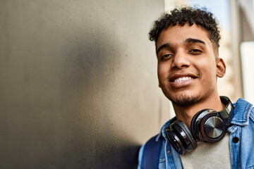 Hispanic young man smiling wearing headphones leaning on the wall