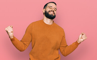 Fototapeta premium Young man with beard wearing casual winter sweater very happy and excited doing winner gesture with arms raised, smiling and screaming for success. celebration concept.