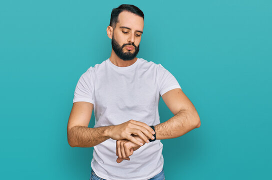Young man with beard wearing casual white t shirt checking the time on wrist watch, relaxed and confident