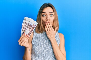 Young caucasian blonde woman holding egyptian pounds banknotes covering mouth with hand, shocked...