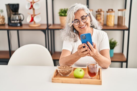 Middle age woman having breakfast using smartphone at home - Powered by Adobe