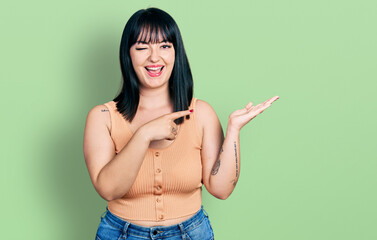 Young hispanic plus size woman pointing open palm with finger winking looking at the camera with...