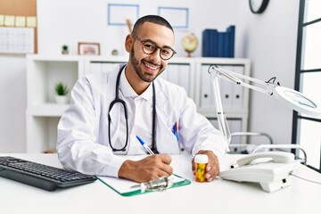 Young hispanic man wearing doctor uniform holding pills at clinic